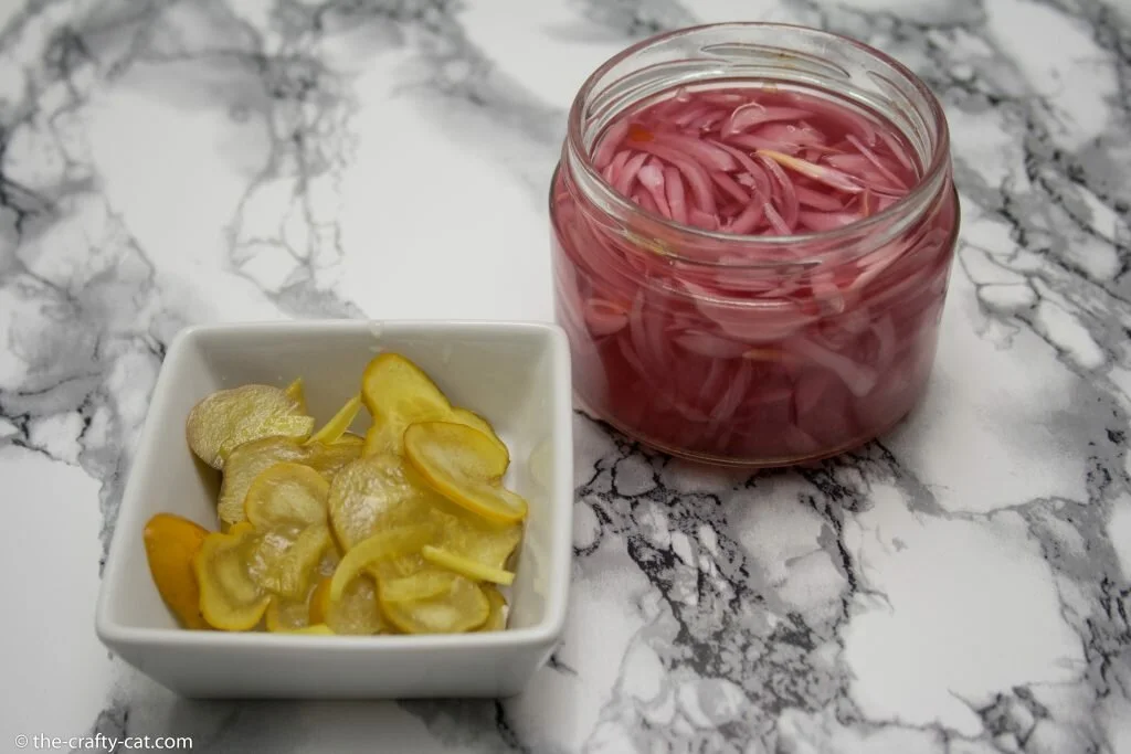 a jar of pickled red onions and a bowl of cucumber pickles with turmeric on a marble table