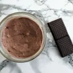 a jar of no-melt chocolate body butter standing on a marble table with a chocolate square next to it