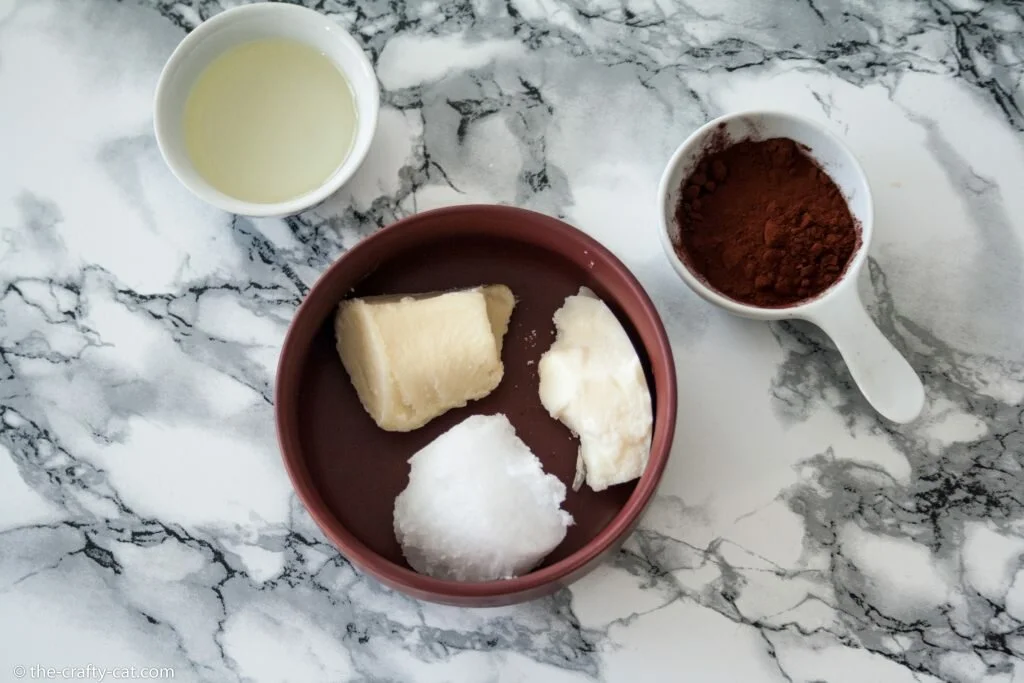 a marble table with a bowl with cocoa butter, shea butter and coconut oil inside as well as two small white bowls containing almond oil and cocoa powder